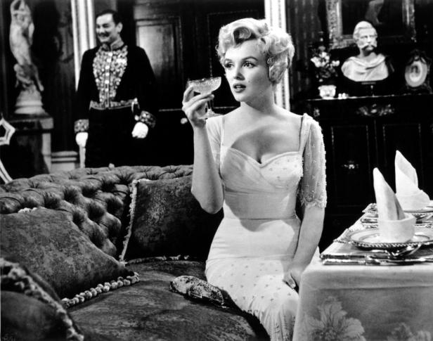 Marilyn Monroe sitzt auf einem Sofa, hat ein weißes Kleid an und eine Champagnerschale in der Hand.