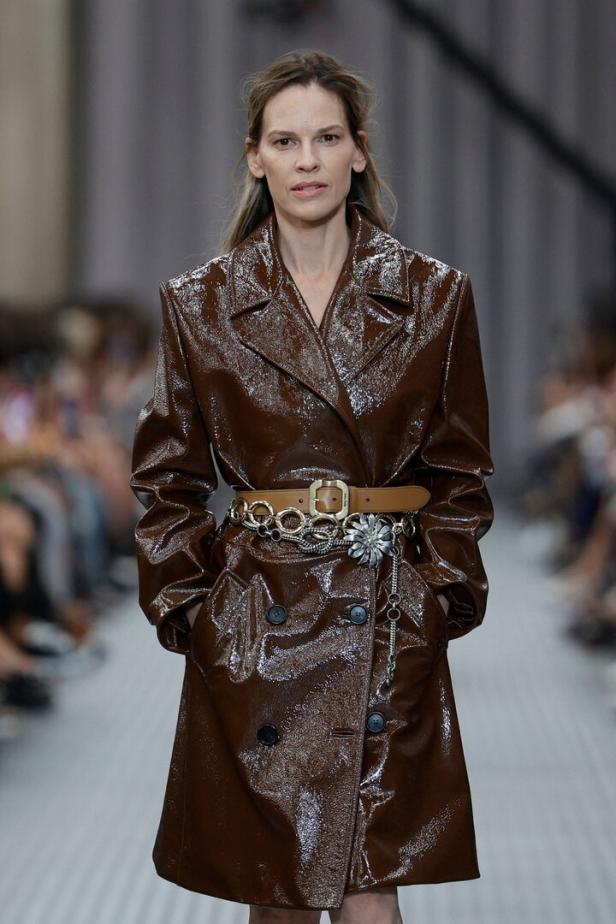 Hilary Swank trägt einen glänzenden, braunen Trenchcoat mit Gürtel auf einem Laufsteg.