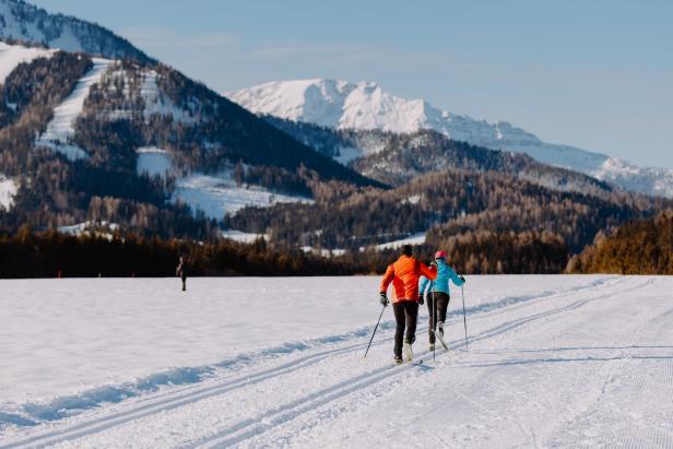 Zwei Personen betreiben Skilanglauf. Im Hintergrund sind hohe, schneebedeckte Berge zu sehen.