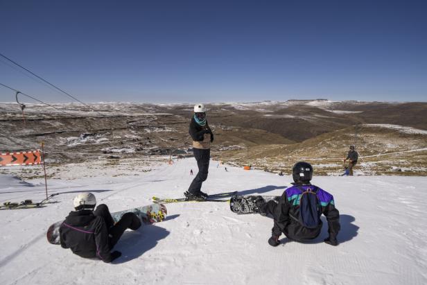 Zwei Sonaboarder sitzen auf der Piste, ein Skifahrer steht. Blick auf eher wenig beschneite Berge