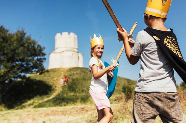 Zwei Kinder spielen Ritter vor einem historischen Gebäude auf einem Feld.