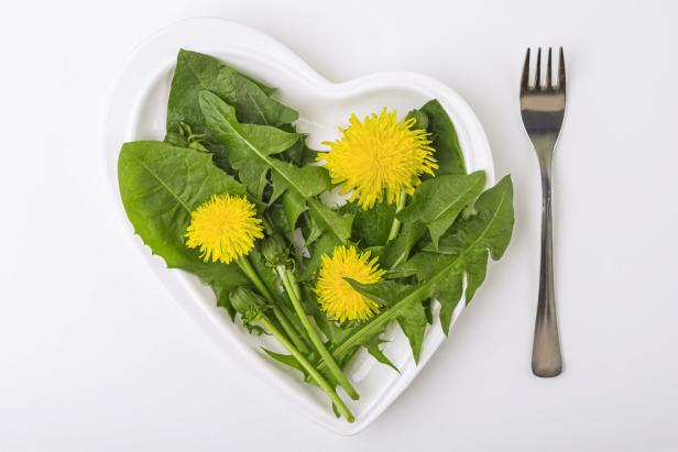 Spring fresh green salad with dandelions and edible flowers on a heart-shaped plate on white background. The concept of diet and healthy eating. Organic and vegetarian food. Copy space for text