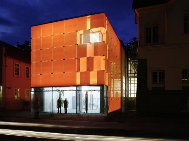 Ein orangenes, kleines, strahlendes Gebäude, in dem sich zwei Personen befinden. Das Erdgeschoss ist aus Glas.