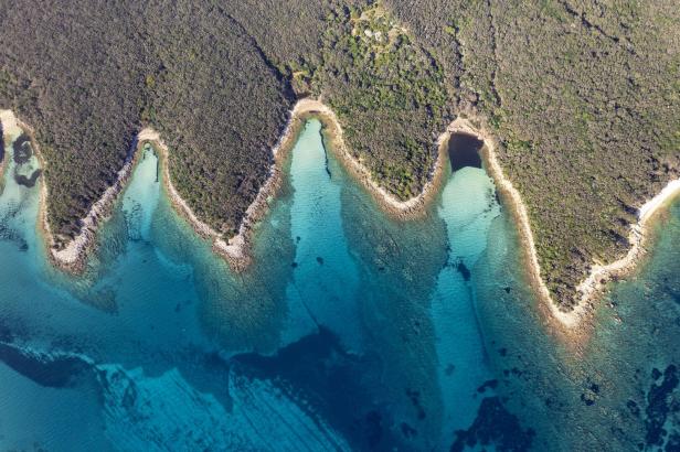 Die Küstenlinie von oben. Das Wasser ist klar, hat unterschiedliche Blauttöne, das Land ist voll von Bäumen.