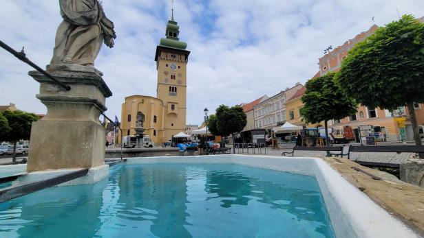 Ein Brunnen auf einem Marktplatz, dahinter ein gelber Turm mit einer grünen Spitze 