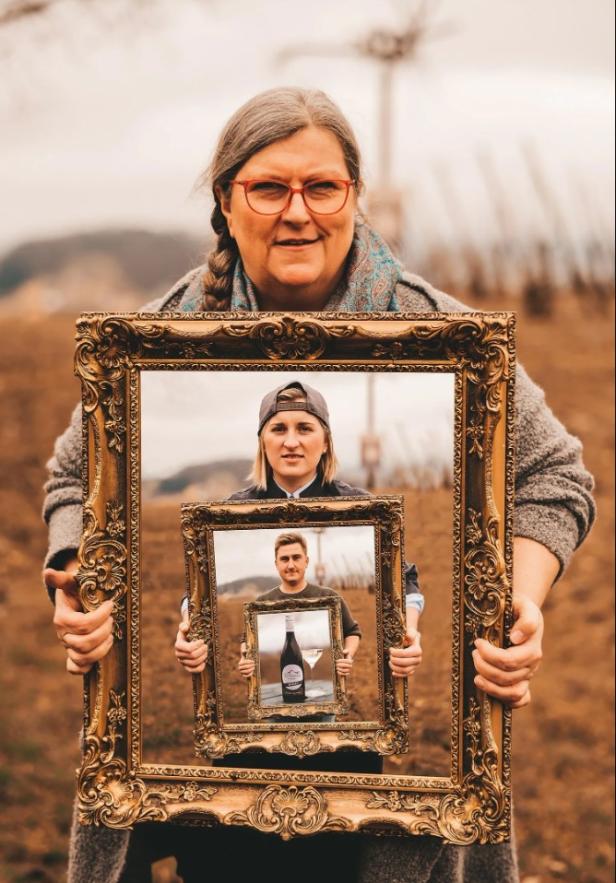 Ilse Jakopé hält einen goldenen Rahmen mit dem Bild ihrer Tochter Lisa-Maria, die wiederum einen Rahmen mit Jakob-Josef hält, der einen Rahmen mit einer Weinflasche zeigt. Das Bild symbolisiert generationenübergreifende Verbundenheit.