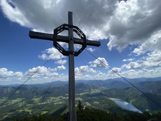 Ein Bild von einem Gipfelkreuz mit Aussicht auf die grüne Berglandschaft und einen See in der Ferne.