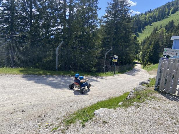 Eine Frau fährt mit einem Kind auf einem Mountaincart auf einer bewaldeten Schotterstraße.
