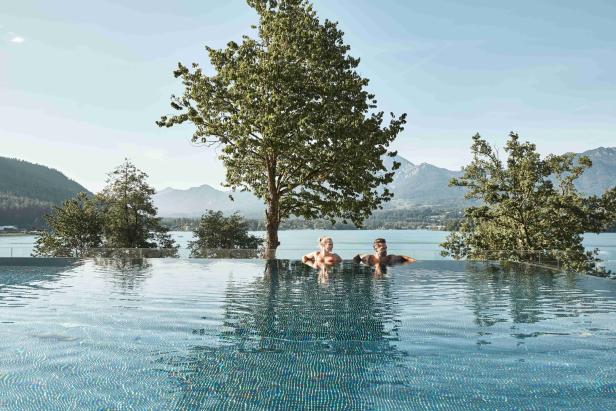 Ein Man und Frau sietzen im Pool, hinter ihnen liegt der Faaker See und dahinter die Berge.