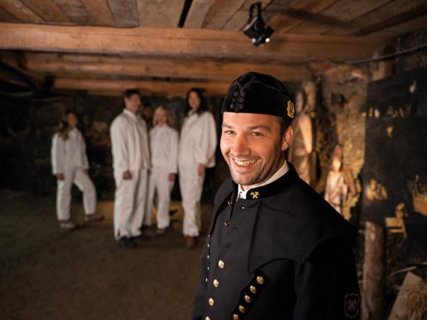 Ein Bergmann in traditioneller Uniform lächelt, im Hintergrund eine Gruppe von Besuchern.