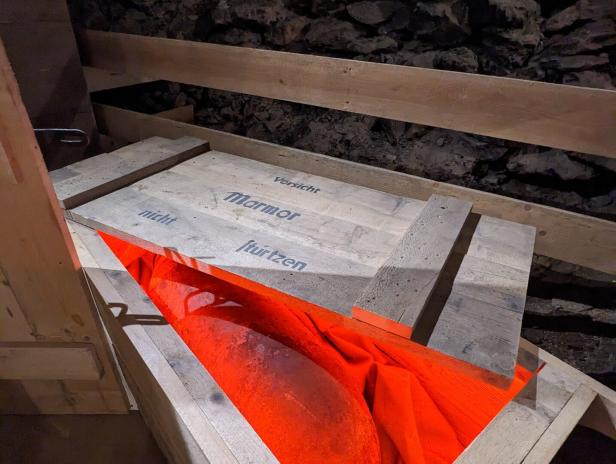 Eine Holzkiste mit der Aufschrift „Vorsicht Marmor, nicht stürzen“ und einem orangefarbenen Tuch darin.