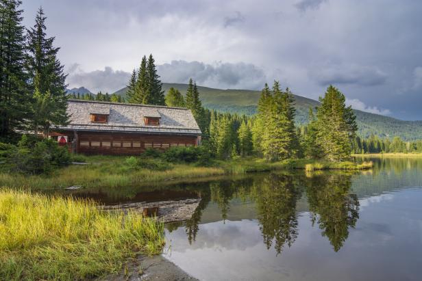 Ein Holzhaus am Ufer eines Sees, umgeben von Bäumen und Bergen.