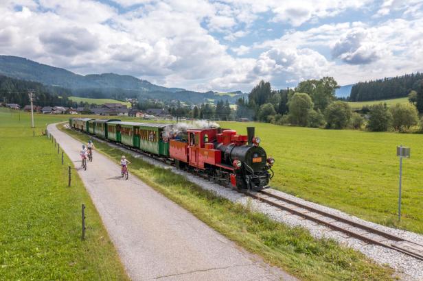 Eine rote Dampflokomotive zieht grüne Waggons durch eine ländliche Gegend, während Kinder auf Fahrrädern nebenher fahren.