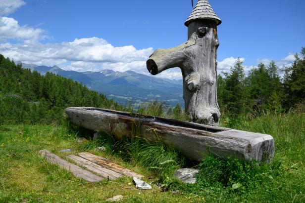 Ein hölzerner Brunnen auf einer Bergwiese mit Blick auf die Alpen.