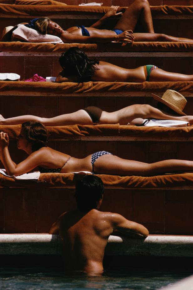 Sonnenbadende Frauen liegen auf Stufen. Ein Mann im Pool sieht zu ihnen hinauf