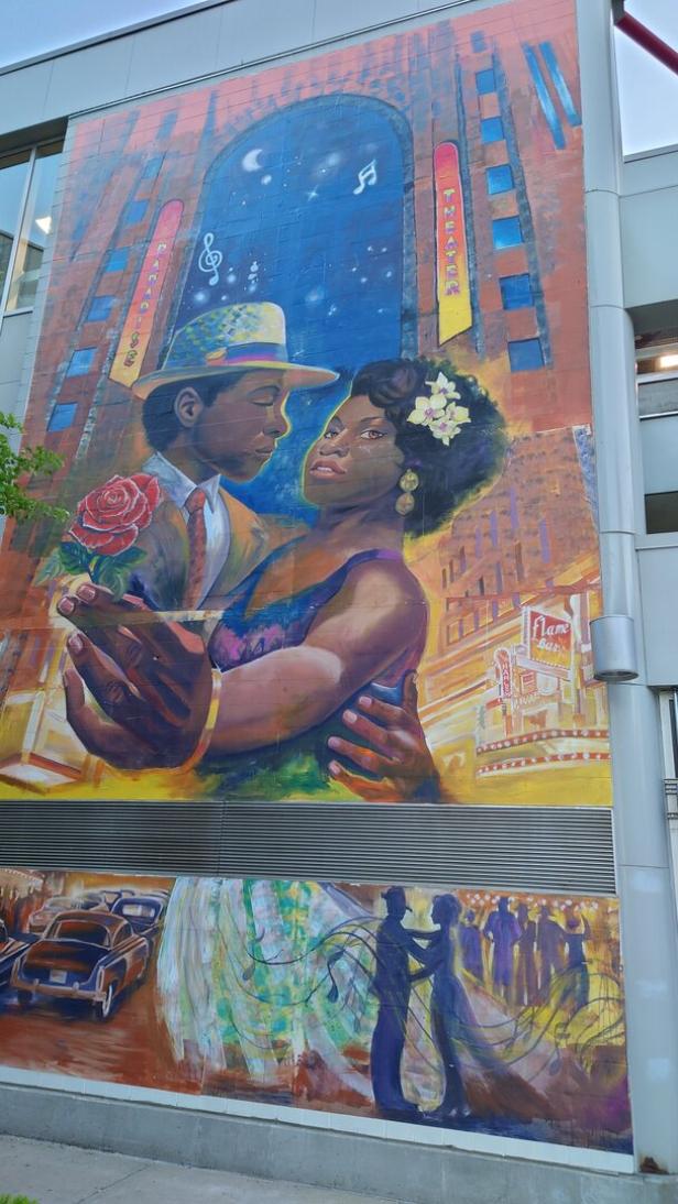 Eine Malerei an einer Hausfassade zeigt ein tanzendes Paar vor dem Hintergrund eines Theaters.