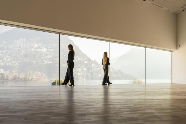 Zwei Frauen stehen in einem Raum mit Panoramafenster und blicken auf einen See und Berge.