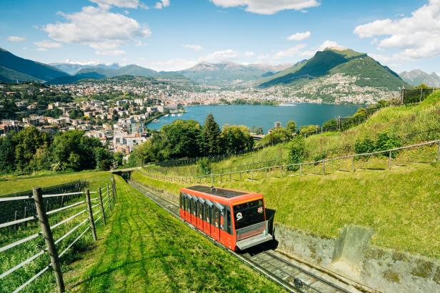 Eine rote Standseilbahn fährt auf einer grünen Bergstrecke mit Blick auf Lugano und den Luganersee.