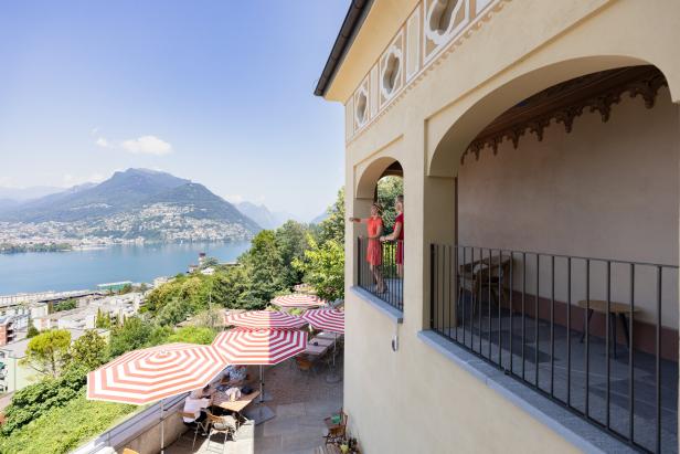 Zwei Frauen stehen auf einem Balkon mit Blick auf den Luganersee und die umliegenden Berge.