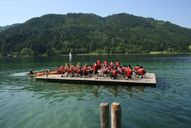 Eine Musikkapelle spielt auf einem Floß auf einem See vor einer bewaldeten Hügellandschaft.