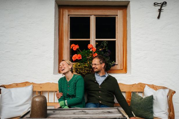 Ein Paar sitzt lachend auf einer Bank vor einem Fenster mit Blumenkasten.