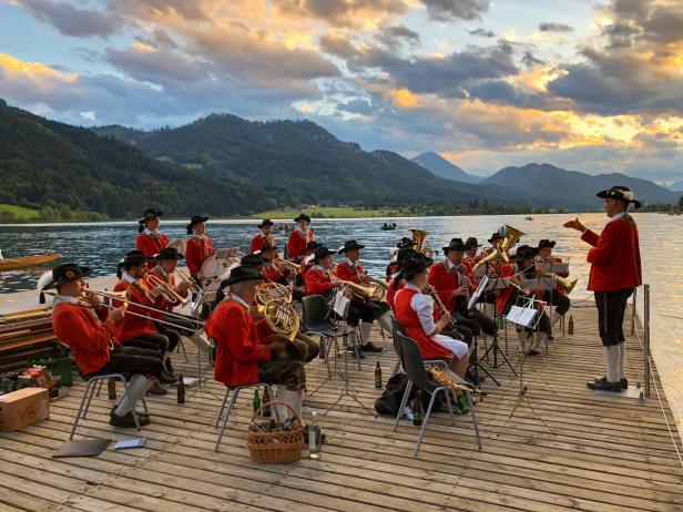 Eine Blaskapelle in Tracht spielt auf einem Steg an einem See vor einer Bergkulisse.