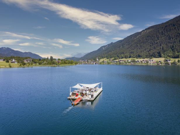 Ein Floß mit Menschen fährt auf einem See vor einer bewaldeten Hügellandschaft.