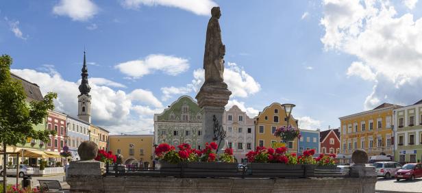 Bunte historische Häuser, eine Statue auf einem Brunnen mit roten Blumen und eine Kirche mit Turm im Hintergrund bei blauem Himmel.