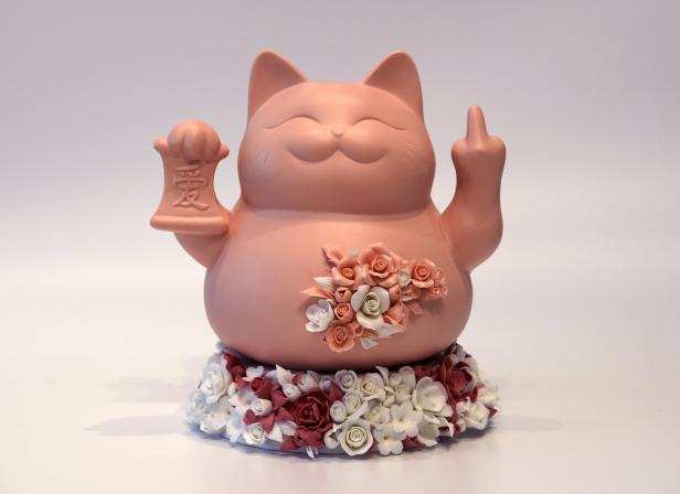 Eine Winkekatze aus Porzellan steht auf Blumen, ist mit Blumen geschmückt, zegigt aber den Mittelfinger