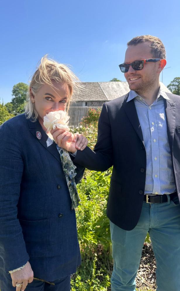 Eine Frau riecht an einer weißen Rose, während ein Mann daneben steht und lächelt.