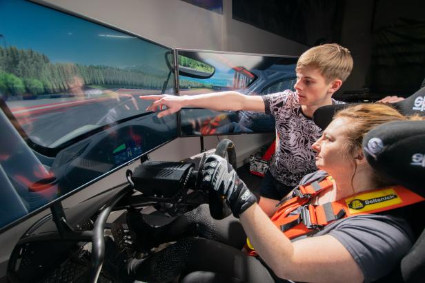 Eine Frau fährt einen Rennsimulator, ein junger Mann zeigt auf den Bildschirm