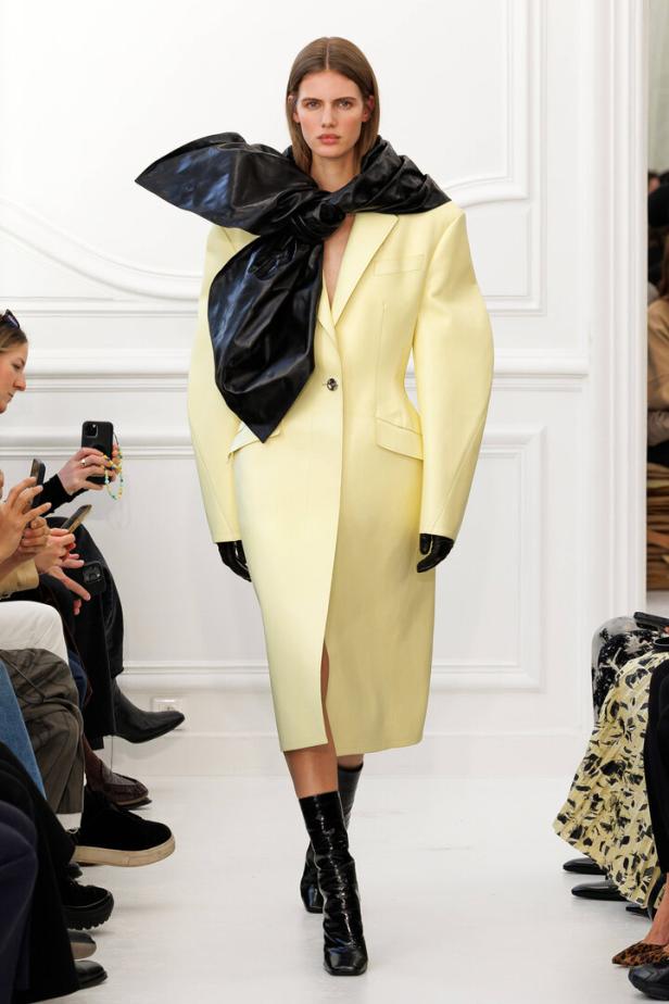 Ein Model präsentiert ein hellgelbes Kleid mit einer großen schwarzen Schleife auf dem Laufsteg.