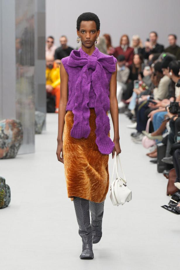 Ein Model auf dem Laufsteg mit einem lila Strickoberteil, einem ockerfarbenen Rock und grauen Stiefeln.