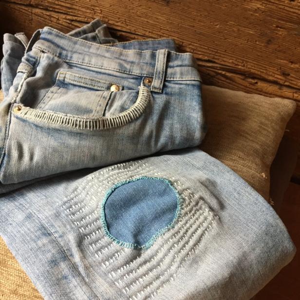 Eine Jeanshose mit geflicktem Loch liegt auf einem Holztisch.