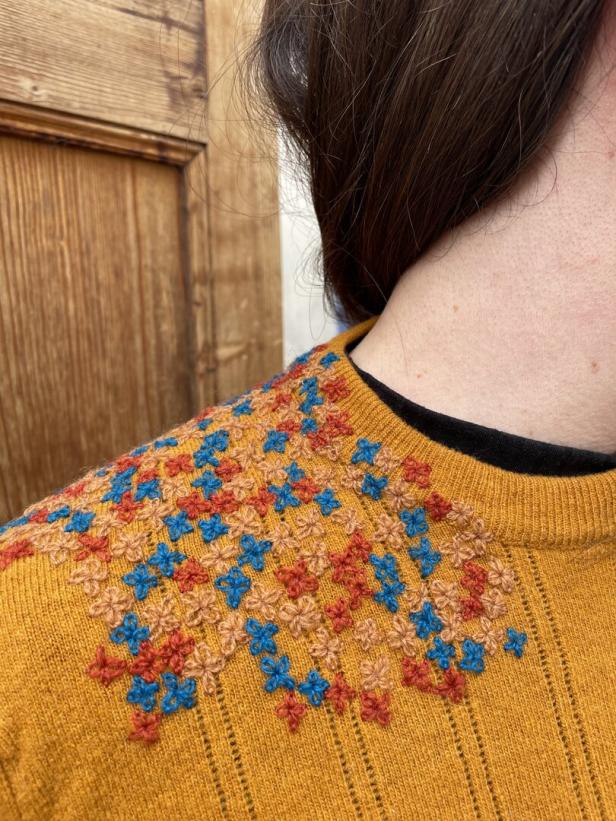 Person zeigt stilvoll geflickten senfgelben Pullover. Es ist nur ein Schulterausschnitt zu sehen.