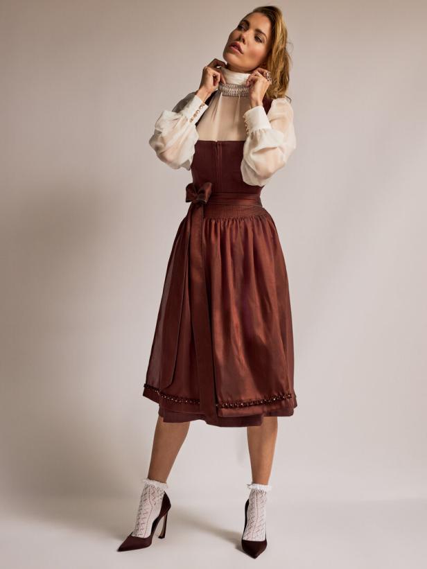 Eine Frau in einem Dirndl mit weißer Bluse und hochhackigen Schuhen posiert vor einem hellen Hintergrund.