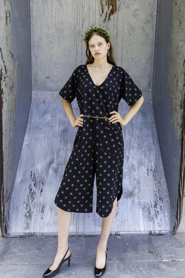 Eine junge Frau posiert in einem gemusterten Jumpsuit und schwarzen Schuhen vor einer grauen Wand.