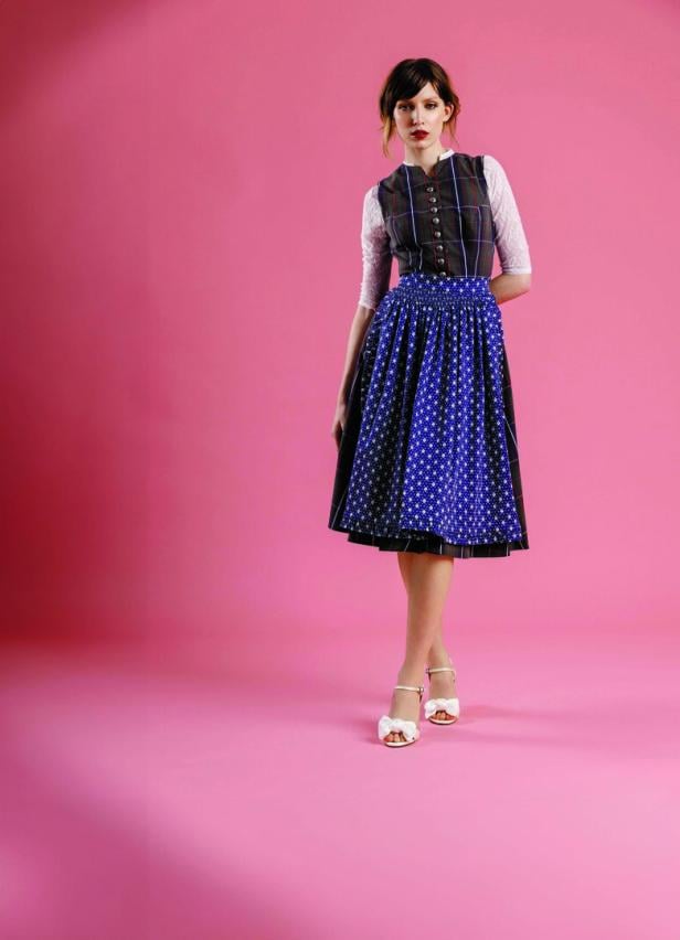 Eine Frau in einem Dirndl mit blauem Rock und karierter Weste vor einem rosa Hintergrund.