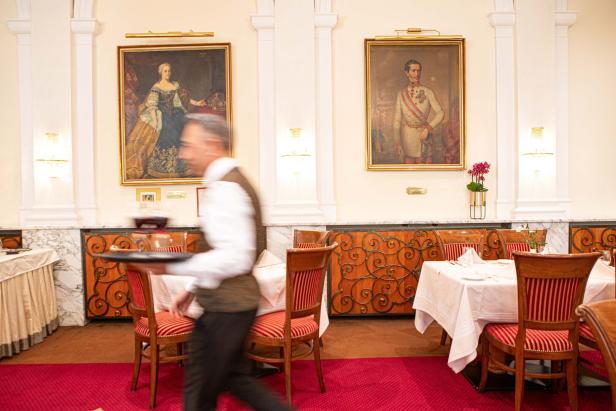Ein Kellner geht an gedeckten Tischen in einem eleganten Restaurant vorbei, an den Wänden hängen Porträts.