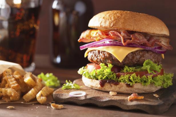Ein Hamburger mit Speck, Käse, Tomaten und Salat, angerichtet auf einem Brett, daneben Pommes Frittes