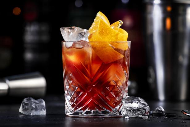 Espresso Negroni alcoholic cocktail drink with dry gin, red vermouth and red bitter, espresso and coffee liqueur, orange slice and ice cubes. Black bar counter background, bar tools, bottles