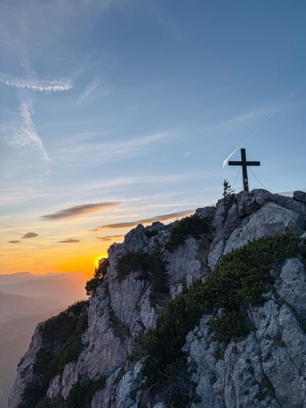 Ein Gipfelkreuz auf einem felsigen Berggrat bei Sonnenuntergang.