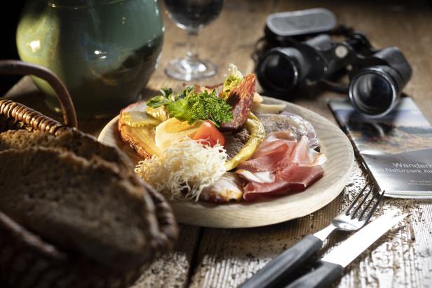 Eine Brotzeitplatte mit Wurst, Schinken und Käse auf einem Holztisch mit Fernglas und Wanderkarte.