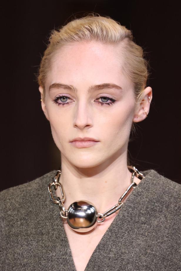 Ein Model mit blasser Haut und blonden Haaren trägt eine auffällige Halskette mit einer silbernen Kugel.