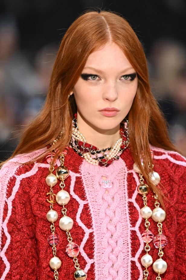 Ein Model mit rotem Haar trägt Chanel-Schmuck und einen roten Pullover auf dem Laufsteg.
