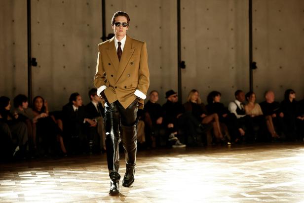 Saint Laurent Menswear Winter 2025 in Paris Model auf dem Laufsteg mit zweireihigem Sakko, das nur unten zugeknöpft ist. Er trägt dazu Overknee-Stiefel aus Leder.