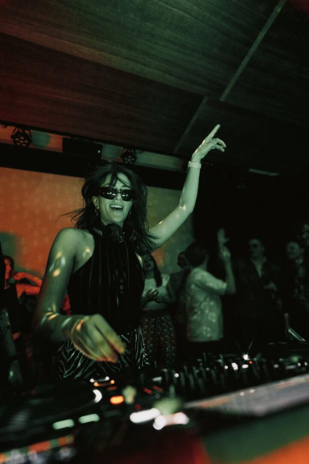 Junge Frau mit Sonnenbrille hinterm DJ-Pult, sie hat den Arm in der Luft.