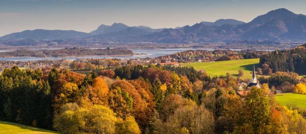 Herbstliche Landschaft mit buntem Laub, grünen Wiesen, einem Dorf mit Kirche und Bergen im Hintergrund unter blauem Himmel.