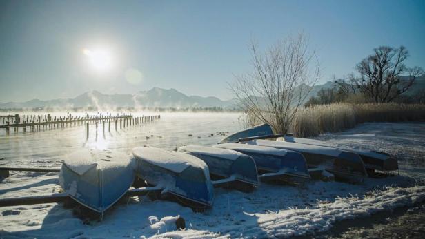 Mehrere mit Schnee bedeckte Boote liegen am Ufer eines Sees, im Hintergrund Berge und ein sonniger Himmel.