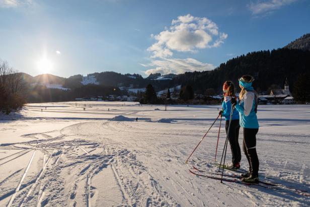 Zwei Skifahrer in blauen Jacken stehen auf einer verschneiten Fläche und blicken auf die Sonne über bewaldeten Hügeln.